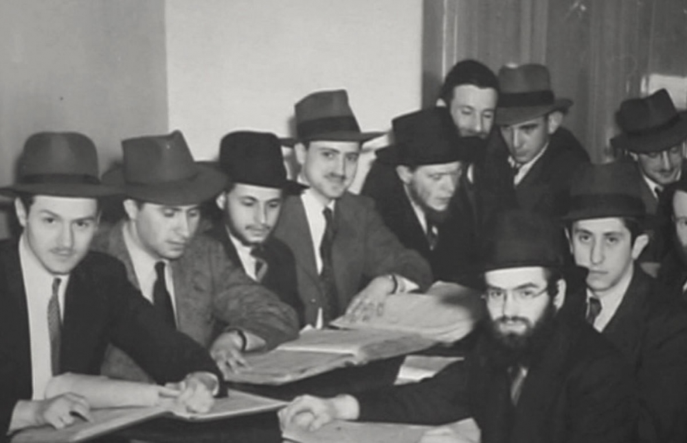 Black-and-white photograph of a group of 10 men sitting together at one table, covered in books and papers. The men wear hats and suits.