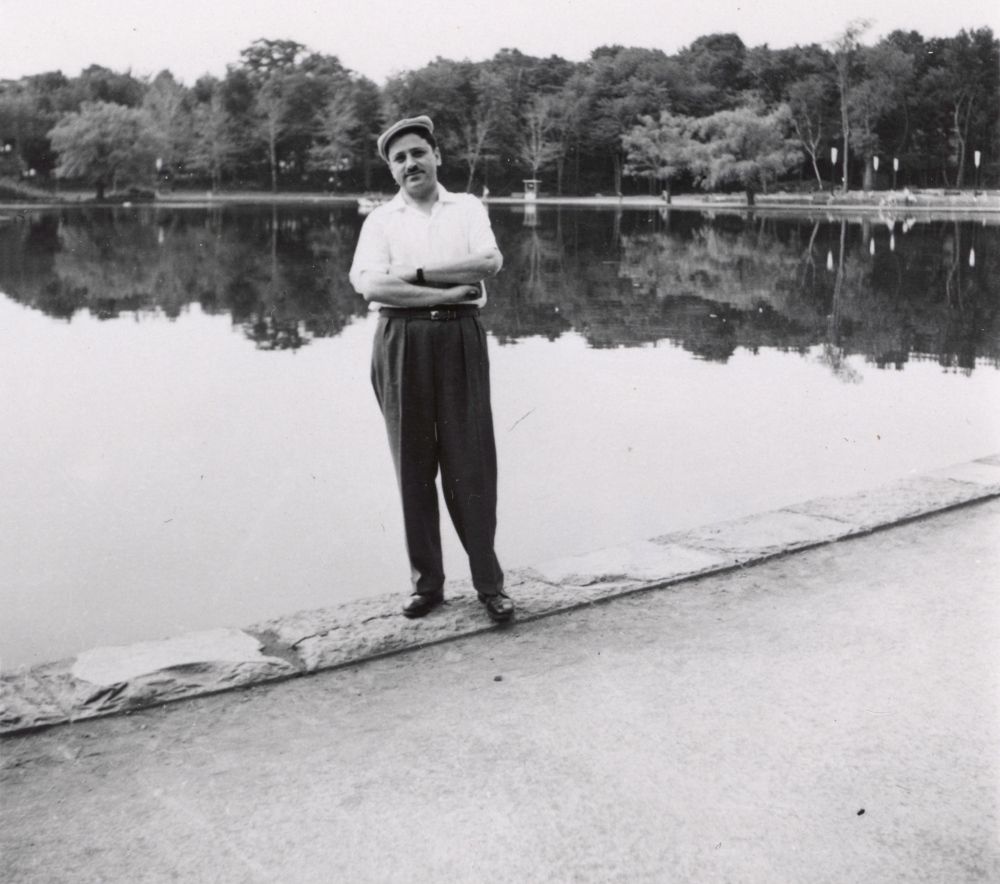 Photo en noir et blanc d'un homme se tenant debout les bras croisés devant un grand étang ou lac. Il porte une chemise à manches courtes, des pantalons noirs et un chapeau. Des arbres entourent le lac en arrière-plan.