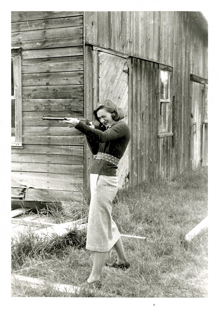 Black-and-white photograph of a woman standing in front of a barn. She holds up a rifle and aims it towards the left of the camera.