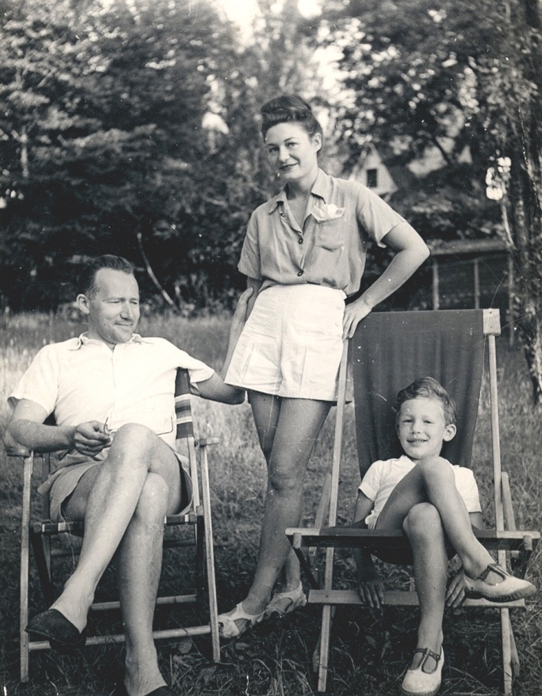 Black-and-white photograph of a woman standing between a man and young boy, who are both sitting with their legs crossed in lawn chairs outside in a field. All three individuals are dressed in shorts and summer clothing.