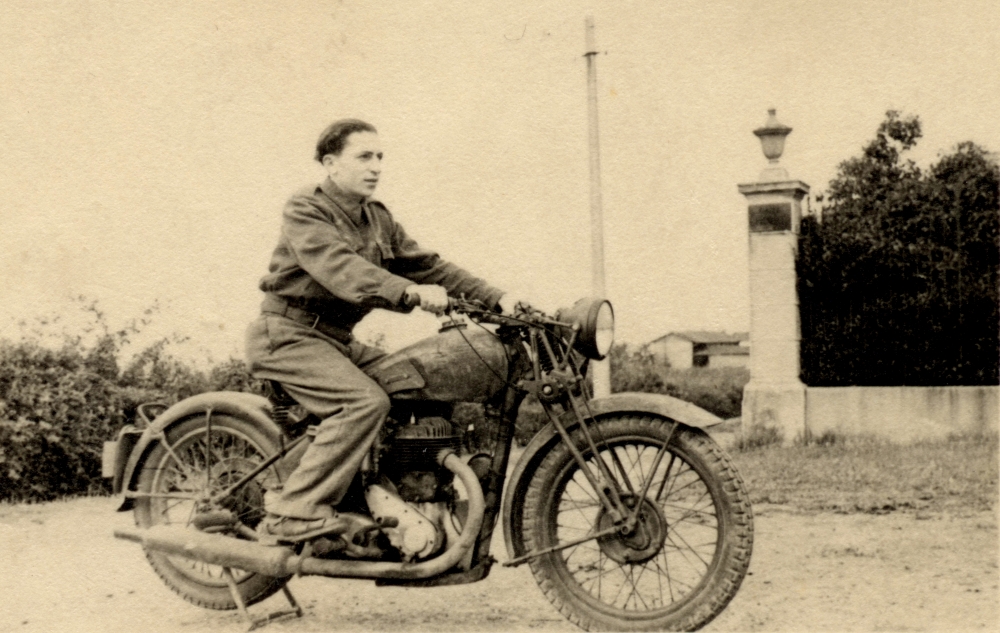 Black-and-white photograph of a young man posing on the back of a motorcycle, looking forwards towards the right of the camera. There is a hedge behind him and a building in the distant background.