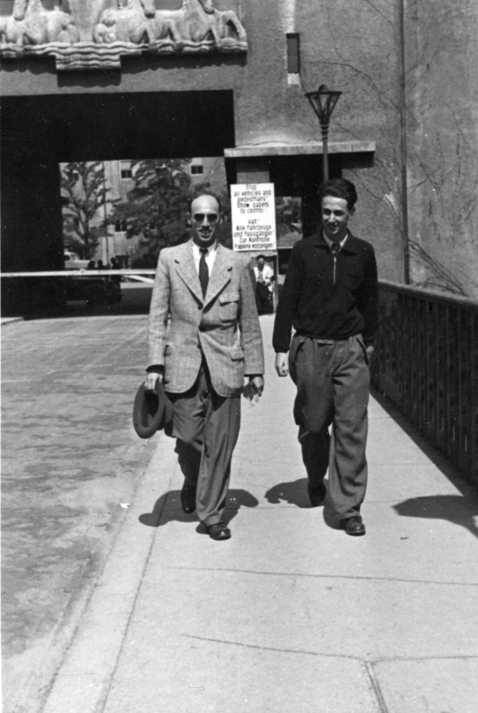 Photo en noir et blanc de deux hommes souriant et marchant vers la caméra sur le trottoir d'un pont. L'homme sur la gauche porte un complet, des verres fumés et tient un chapeau dans sa main droite.