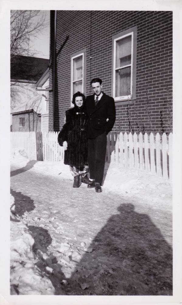 Photo en noir et blanc d'une femme et d'un homme debout ensemble à l'extérieur sur une allée enneigée devant une clôture blanche. La femme porte un manteau de fourrure et l'homme un caban de couleur foncée.