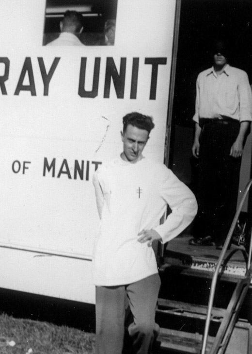 Black-and-white photograph of a man, standing with a hand on his left hip, in front of a large white truck that reads “X-RAY UNIT” on its side. A man stands behind him inside the entrance of the truck with his face covered by shadow.