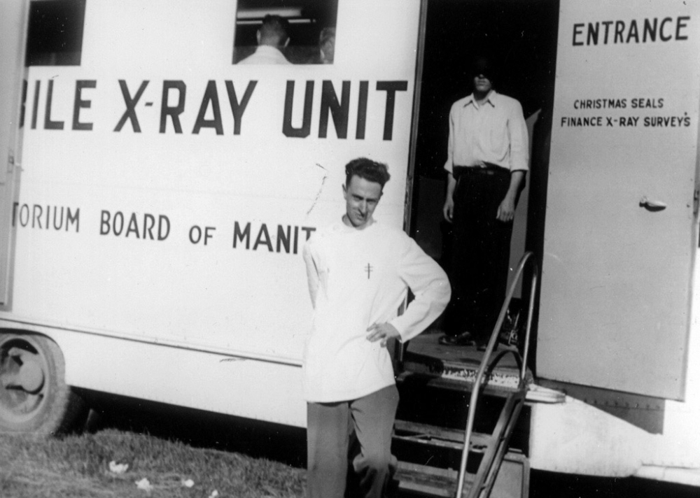 Black-and-white photograph of a man, standing with a hand on his left hip, in front of a large white truck that reads “X-RAY UNIT” on its side. A man stands behind him inside the entrance of the truck with his face covered by shadow.