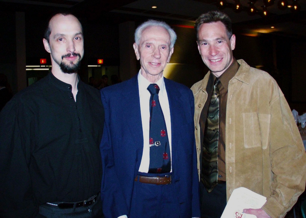 Colour photograph of three men, shown from the waist upwards, smiling at the camera. The elderly man in the middle wears a blue suit and tie, the man on the left wears a black shirt and has facial hair. The man on the right wears a camel jacket and brown shirt with tie.