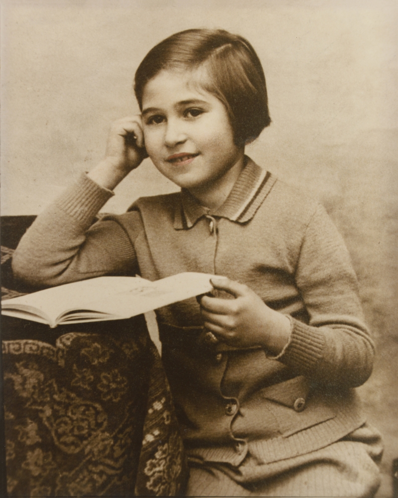 Sepia-tone photograph of a young girl, sitting at a desk with a tablecloth. The girl rests an open book on the desk with one hand, and leans on her other elbow in a pensive pose. Light pink shading has been added to her lips.