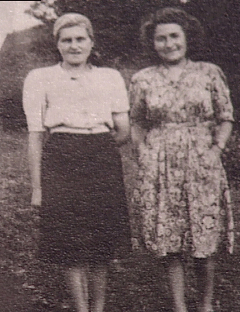 Photo en noir et blanc de deux jeunes femmes debout à l'extérieur et souriantes. Une des femmes porte une robe et l'autre une blouse et une jupe.