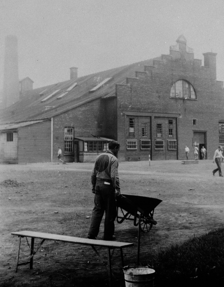 Black-and-white photograph of a man, with his back to the camera, pushing a wheelbarrow in a yard. There is a bench in the foreground, and a building in the far background. The man wears a uniform with a big circle on his back.