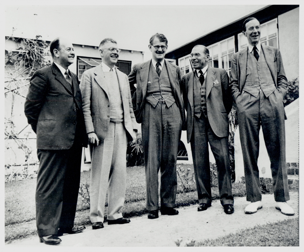 Black-and-white photograph of five men, wearing suits, standing outside in a row and smiling or laughing.
