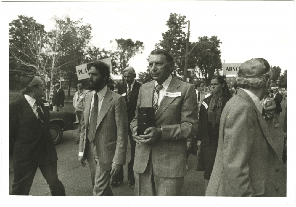Photo en noir et blanc de quatre hommes menant une marche à l'extérieur. L'homme au centre transporte une boîte de forme rectangulaire. Un groupe de personnes les suit en arrière-plan.