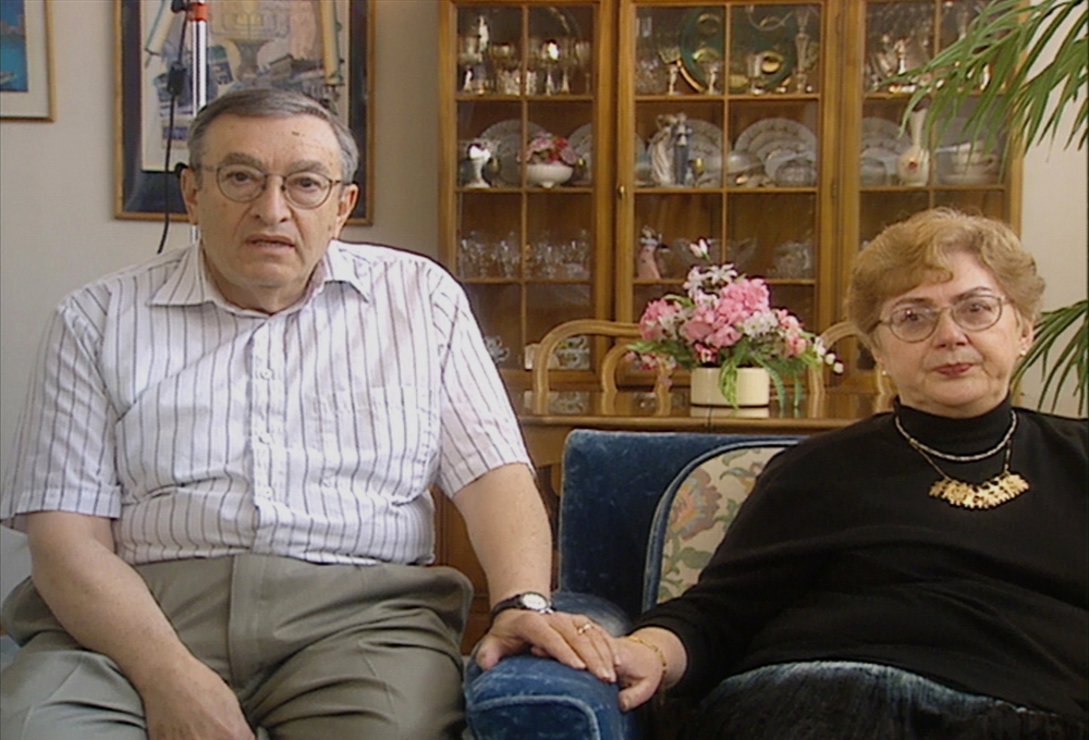 Colour photograph of a elderly man and woman sitting together indoors, holding hands. The woman sits on a blue arm chair. There is a display cabinet containing dishes and chinaware in the background.