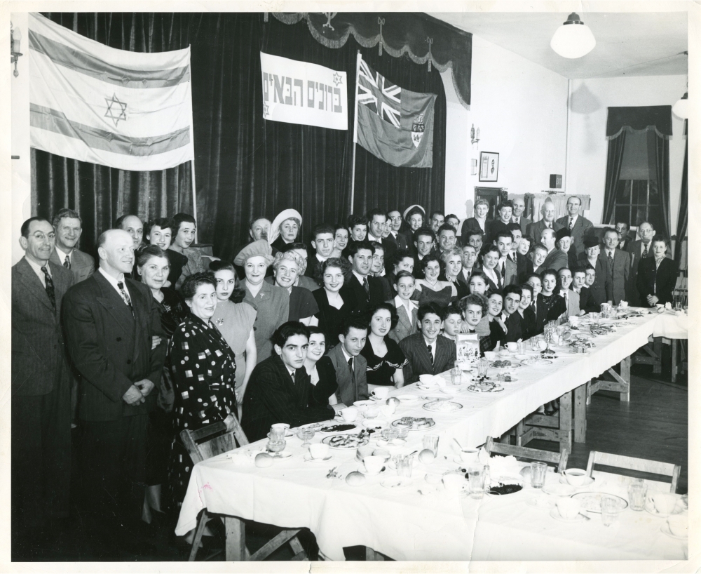 Black-and-white photograph of a large group of approximately 70 people smiling for a group photograph in a room. The group poses behind a large table with food and dishes on it, and there are two flags hung on curtains in the background.