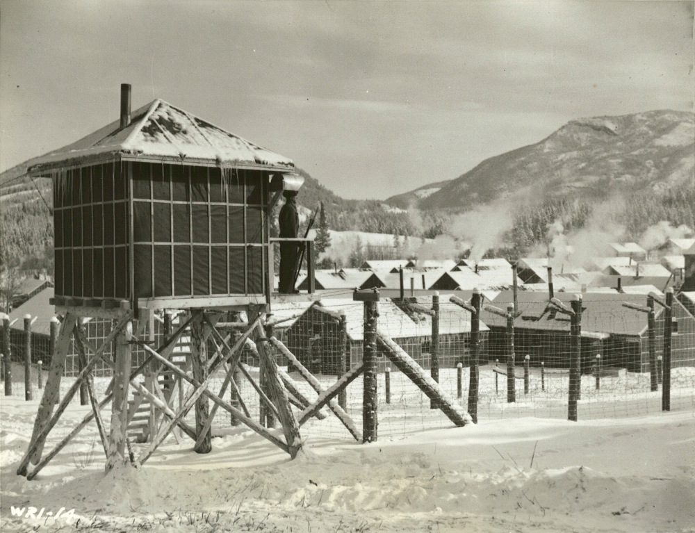 Black-and-white photograph of a guard standing at attention in his barracks, surrounded by barbed wire. Rows of buildings are in the background, with snow-covered hills in the distance. There is snow on the ground.