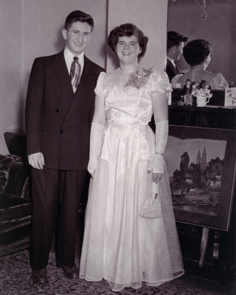 Photo en noir et blanc d'un jeune homme et d'une jeune femme se tenant debout à l'intérieur et souriant ensemble devant un mirroir. La femme porte une longue robe formelle de couleur claire ainsi qu'un petit sac à main et l'homme porte un complet.