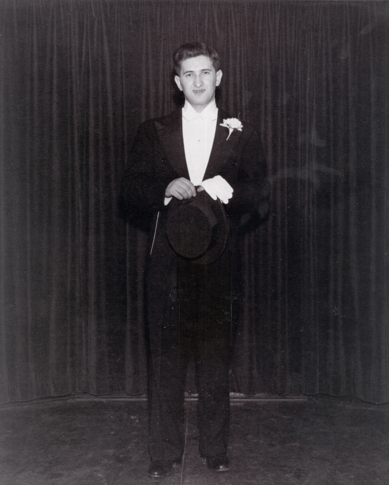 Photo en noir et blanc d'un jeune homme portant un tuxedo , se tenant devant un rideau de couleur foncée.