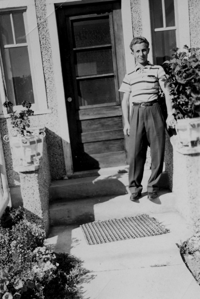 Black-and-white photograph of a young man standing outdoors, on the first step of an entrance to a house. He wears a short-sleeved striped shirt and trousers.