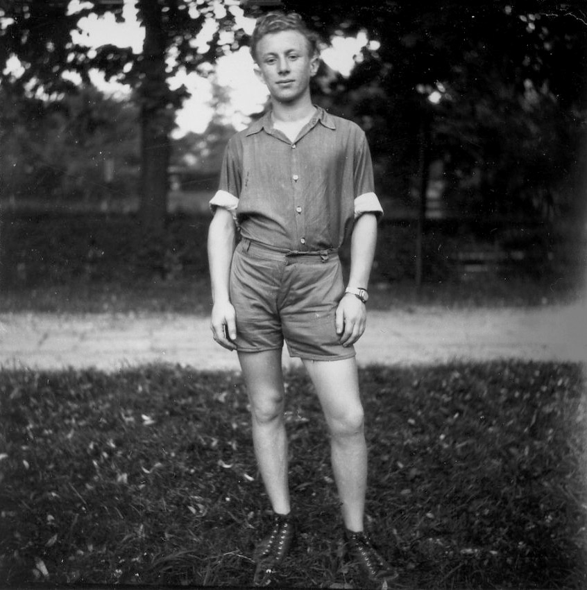 Black-and-white photograph of a young man standing outdoors on a field. It appears to be summer because the young man wears a short-sleeve rolled up shirt and shorts.