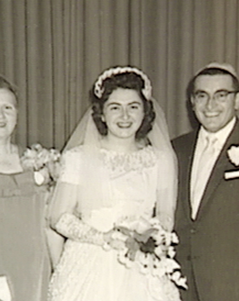 Photo en noir et blanc d'une femme debout et souriant à la caméra pendant son mariage. Elle porte une longue robe blanche, un voile et tient un bouquet de fleurs.