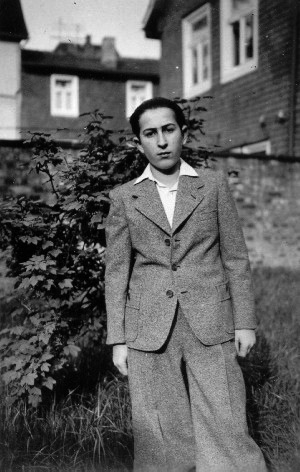 Photo en noir et blanc d'un jeune homme debout à l'extérieur dans ce qui semble être une cours arrière avec une clôture et un arbuste derrière lui. Il porte un complet et une chemise à col, ses cheveux sont coiffés vers l'arrière.