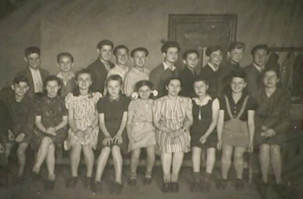 Black-and-white photograph of a classroom, grouped together in two rows of about 10-12 children each. The girls wear dresses, sitting in the front row and the boys stand in the back with collared shirts.