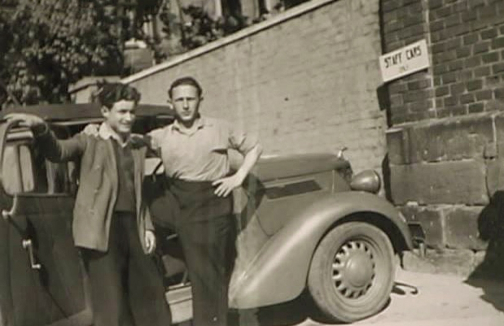 Photo en noir et blanc de deux jeunes hommes debout devant une voiture ancienne à l'extérieur. Un des hommes entoure l'autre de son bras. Il y a un grand mur de brique en arrière-plan.