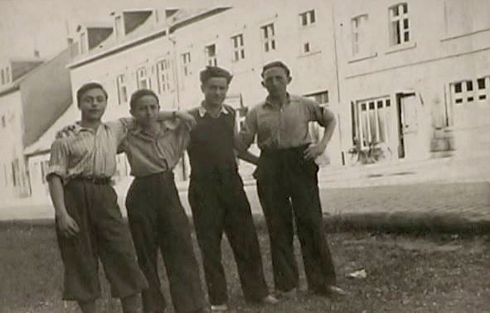 Photo en noir et blanc d'un groupe de quatre garçons se tenant bras-dessus bras-dessous sur la partie ombragée d'une route. Il y a un grand édifice de l'autre côté de la rue.