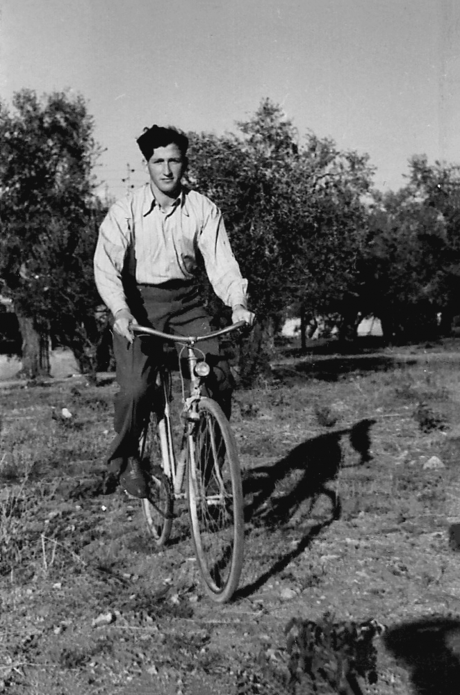 Black-and-white photograph of a young man riding a bicycle in a field. He wears dark trousers and a light collared shirt. There are trees in the background.