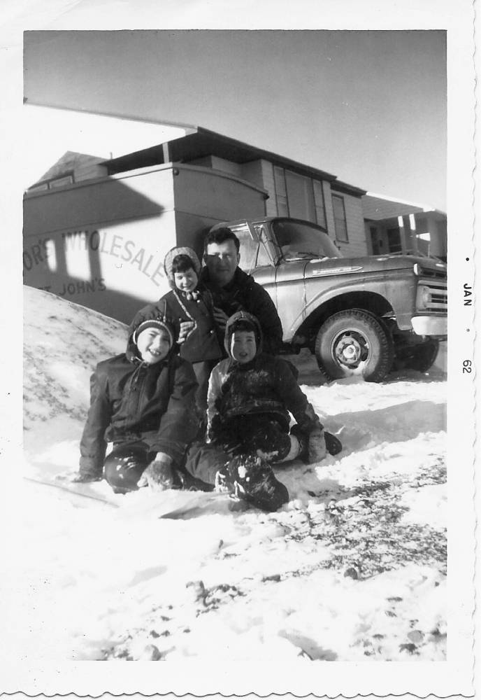 Black-and-white photograph of man with his three young children, sitting in the snow outdoors. They smile at the camera, grouped together in front of a house with a truck in the background.