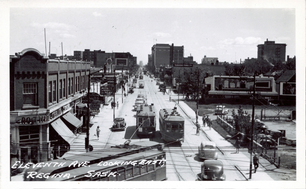 Black-and-white photograph of a street scene, taken from a high vantage point. The large street has pedestrians on the sidewalks, storefronts on either side, and three streetcars driving along in the middle of the road.