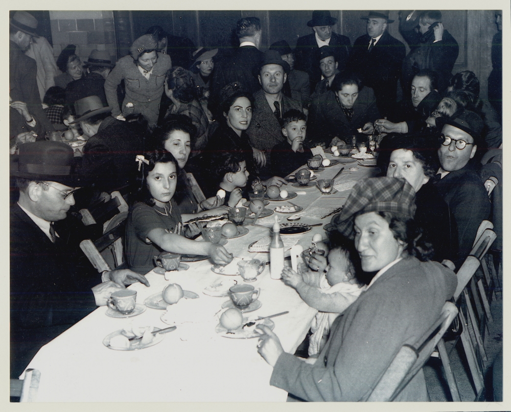 Photo en noir et blanc d'un grand groupe de personnes assis ensemble à longue table, regardant vers la caméra. La table est remplie de plats et de nourriture. Il y a plusieurs personnes dans la pièce derrière la table.