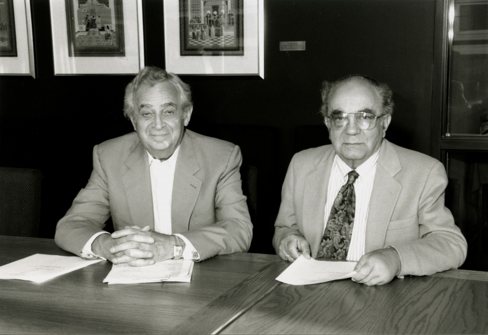 Black-and-white photograph of two elderly men sitting at a table with papers in front of them. The men wear suit jackets, one them wearing a tie, and there are three framed artworks hung on a wall in the background.