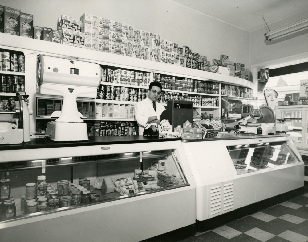 Photo en noir et blanc d'un homme se tenant derrière une caisse enregistreuse dans une grand épicerie. Il se tient devant une étagère remplie de produits et il y a un grand comptoir de présentation devant lui.