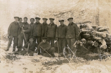 Black-and-white photograph of a group of eleven men posing together for a photograph in the woods. Some of the men are holding axes. They are grouped together in front of a pile of wood, wearing matching outdoor uniforms.