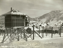Une photographie en noir et blanc d'un garde qui se tient dans sa caserne entourée de barbelés. Les rangées de bâtiments sont en arrière-plan, avec des collines couvertes de neige au loin. Il y a de la neige au sol.
