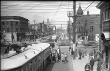 Black-and-white photograph of a busy street intersection. Pedestrians board a streetcar in the bottom-left corner, and cable wires criss cross the air, visible in the top section of the image.