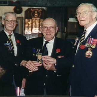 Colour photograph of three elderly men standing together and holding a military medal. The men wear suit jackets adorned in pins and medals.