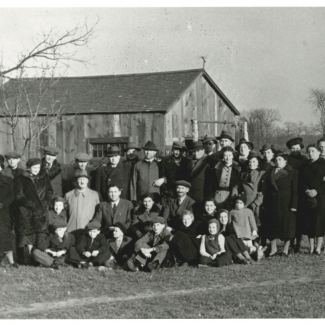 Black-and-white photograph of large group of about 35 men, women, and children, dressed in winter coats and hats, grouped together outdoors on a lawn. There is a barn in the background behind the group.