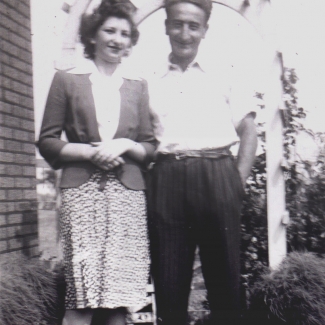 Photo en noir et blanc d'un homme et une femme se tenant ensemble à l'extérieur sous une tonnelle, souriant à la caméra. L'homme a les mains dans les poches.