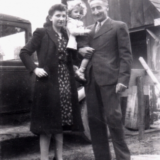 Photo en noir et blanc d'un homme et d'une femme avec un jeune enfant au cheveux blonds bouclés. Ils sont ensemble à l'extérieur devant un camion et une grange en arrière-plan.