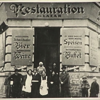 Photo en noir et blanc d'un groupe d'environ huit personnes se tenant devant l'entrée d'un restaurant. L'édifice a une grande affiche de chaque côté de la porte, rédigées en allemand. Deux jeunes enfants regardent le groupe d'un des deux côtés.
