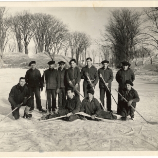 Photo en noir et blanc d'une équipe de hockey de 11 hommes, posant ensemble avec leur bâton de hockey sur la glace d'une patinoire extérieure. Des arbres sont visibles en arrière-plan.