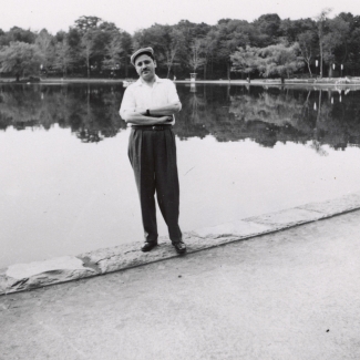 Black-and-white photograph of a man standing with his arms crossed, in front of a large pond or lake. He wears a short-sleeved shirt, dark trousers, and a cap. Trees line the lake in the background.
