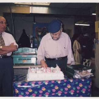 Colour photograph of an elderly man with a beard and cap, cutting into a large cake while two men stand on either side watching him.