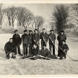 Black-and-white photograph of a hockey team of 11 men, posing together with hockey sticks on an ice rink outdoors. Trees are visible in the background.