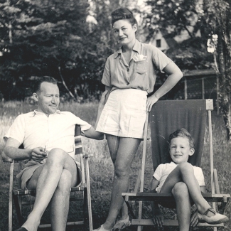 Black-and-white photograph of a woman standing between a man and young boy, who are both sitting with their legs crossed in lawn chairs outside in a field. All three individuals are dressed in shorts and summer clothing.