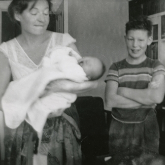 Black-and-white photograph of a young boy standing with his arms crossed and smiling beside a woman who holds a crying baby wrapped in a blanket.