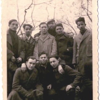 Photo en noir et blanc d'un groupe de neuf jeunes hommes se tenant ensemble bras-dessus bras-dessous. Ils portent des manteaux et il y a des arbres nus au-dessus d'eux en arrière-plan.