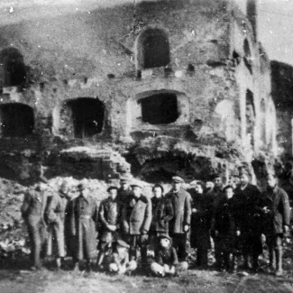 Black-and-white grainy photograph of a group of 15 people posing together outdoors in a line, 2 children sitting on the ground, in front of the ruins of a building. The building is missing a roof and has several window frames that no longer have any windows.