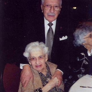 Colour photograph of two elderly people smiling at the camera. The man stands behind the woman, who is sitting at a chair, with his hands on her shoulders. She holds one of his hands.